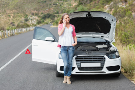 Woman leaning against sedan hood open on mountain road placing red triangle calling on smartphone. Automobile, roadside assistance, outdoor, adventure, lifestyle, travel, ruggedの写真素材