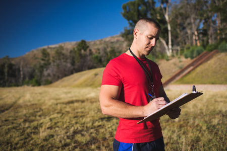Man standing in grassy field by wooden stairway writing on clipboard with whistle and pen. Landscape, fitness, outdoor, wellness, motivation, activity, naturalの写真素材