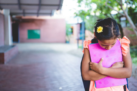 Girl standing clutching pink binder, wearing black backpack on walkway under corridor with trees. Child, student, campus, outdoor, education, youthful, natureの写真素材