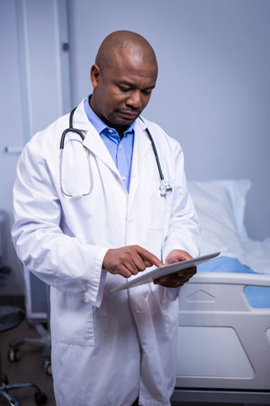 African American male physician reviewing tablet in hospital room with stethoscope and bed linens. Medical, healthcare, professional, clinical, diagnostic, patient care, technologyの写真素材