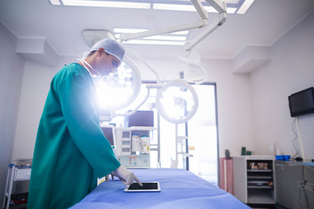 Male surgeon wearing green gown under surgical lights in operating room using tablet, copy space. Medical, surgical, healthcare, hospital, professional, blue, equipmentの写真素材