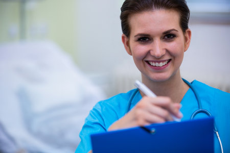 Female nurse smiling filling out chart by bed in ward with clipboard, stethoscope, copy space. Healthcare, professional, medical, clinical, caregiver, wellness, documentationの写真素材