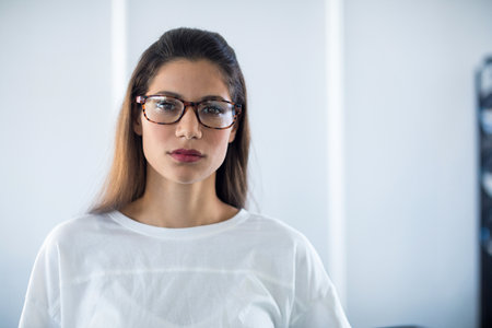 Woman standing against off-white wall in room wearing tortoiseshell eyeglasses and white shirt....