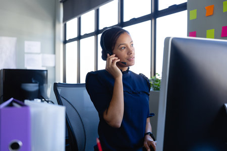 African American woman using computer and headset while sorting binder files at modern office desk....