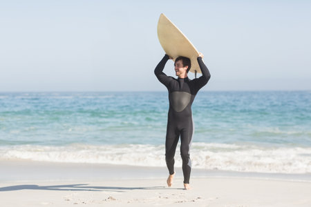 Male surfer wearing wetsuit lifting surfboard overhead on sandy shore under clear sky, copy space. Adventure, sport, coastline, leisure, outdoor, energetic, thrillの写真素材