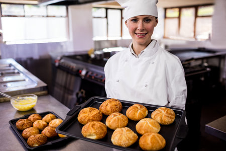 Female chef in chef coat and hat, holding baking tray with golden rolls in restaurant kitchen. Bakery, culinary, artisan, rustic, gastronomic, gourmet, hospitalityの写真素材