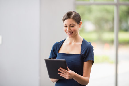 Woman standing in office lobby wearing navy blue dress holding black tablet and scrolling by window. Professional, modern, sleek, corporate, technology, lifestyle, businessの写真素材