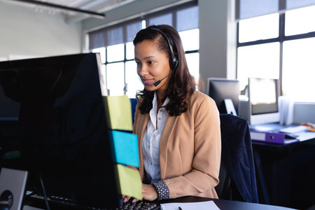 African American woman sitting at office desk typing on keyboard with headset near sticky notes. Professional, workspace, modern, communication, corporate, productivity, ergonomicの写真素材