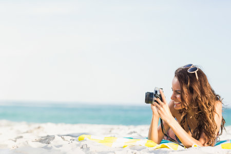 Woman in bikini lying on beach towel shooting blue ocean holding camera and sunglasses, copy space. Vacation, leisure, serenity, outdoor, lifestyle, adventure, minimalistの写真素材
