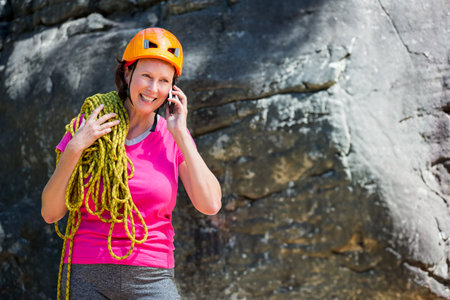 Woman standing at base of rock wall wearing helmet and rope, talking on smartphone, copy space. Adventure, outdoor, rugged, resilience, exploration, athletic, natureの写真素材