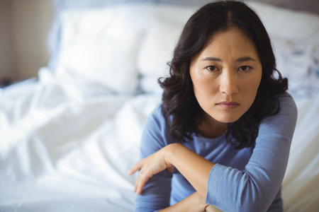 Asian woman sitting on bed with white pillows and sheets folding arms and looking serious. Serenity,...