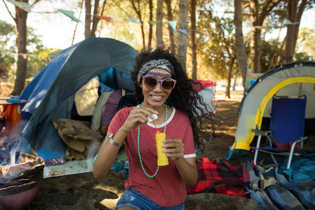 African American woman sitting on blanket blowing bubbles with bubble wand at campsite with tents. Outdoor, leisure, rustic, playful, nature, adventure, vibrantの写真素材
