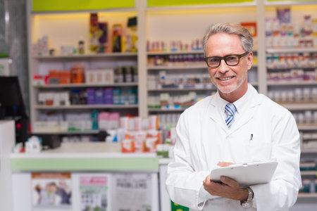 Male pharmacist wearing lab coat standing at pharmacy counter holding clipboard and checking stock. Healthcare, professionalism, medical, consultation, retail, wellness, diagnosticの写真素材