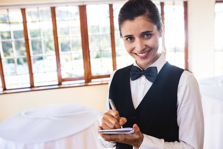 Female event server smiling holding notepad, pen next to grid windows, round tables, copy space. Hospitality, service, professional, elegant, choreography, interior, event planningの写真素材