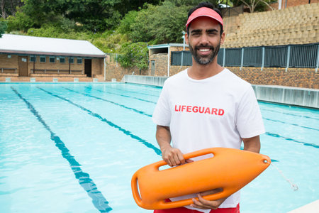 Male lifeguard wearing uniform holding orange rescue buoy at pool edge with lane lines, copy space. Rescue, safety, aquatic, vigilance, sport, outdoor, recreationalの写真素材