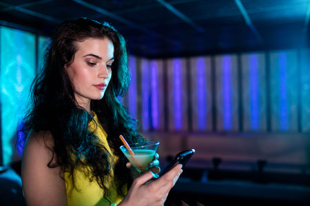 Woman wearing yellow dress holding smartphone and martini glass near bench in lounge, copy space. Luxury, nightlife, elegant, ambient, stylish, modernism, socializingの写真素材