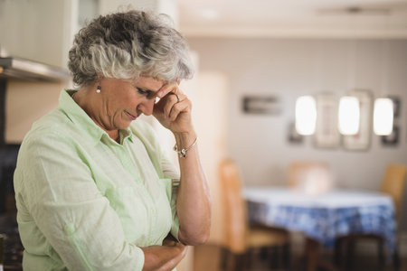 Senior woman standing in home kitchen near oven pressing finger to temple on countertop, copy space. Casual, cozy, contemporary, homemaking, domestic, reflective, sereneの写真素材
