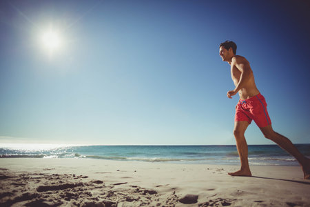 Shirtless runner jogging on wet sand under bright sun with ocean waves rolling, copy space. Athletic, leisure, outdoor, natural, energetic, vitality, freedomの写真素材