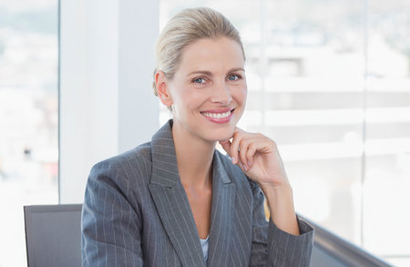 Smiling mature adult woman wearing gray pinstripe blazer resting chin on hand by windows in office. Professional, corporate, modern, confident, optimistic, executive, workspaceの写真素材