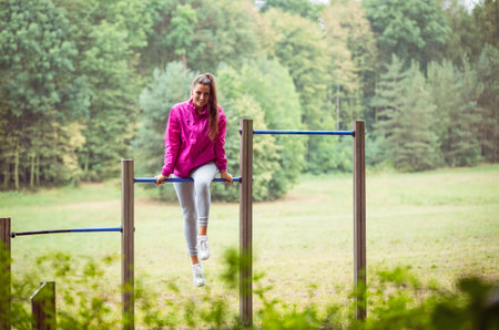 Woman in pink windbreaker performing hanging leg raises on metal exercise frame in park clearing....