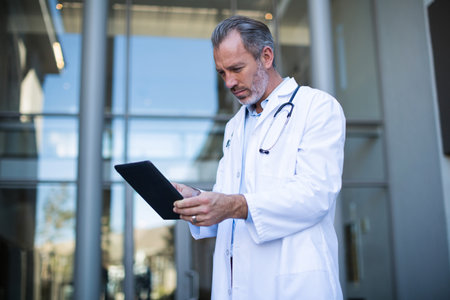 Senior male physician standing at clinic door reviewing tablet and wearing stethoscope, copy space....