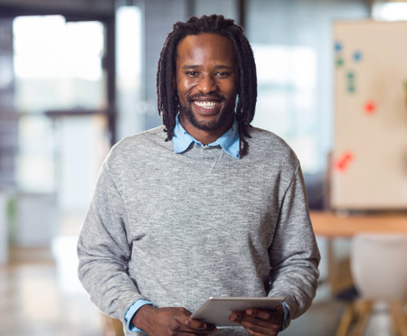 Mid-adult African American man smiling and holding tablet computer near whiteboard in modern office. Professional, innovation, collaboration, technology, workspace, productivity, businessの写真素材