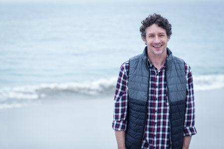Man in his 40s wearing quilted vest standing on beach with waves rolling, copy space. Coastline,...