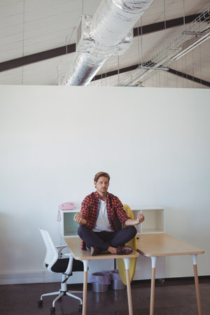 Man in his thirties meditating on light wood desk in office with pastel pink telephone. Concentration, mindfulness, tranquility, corporate, modern, interior, wellnessの写真素材