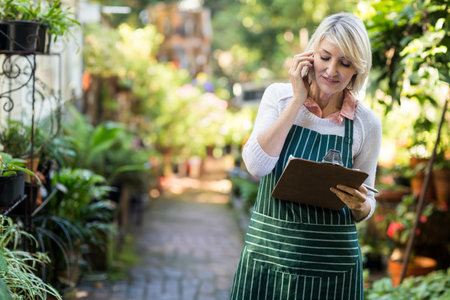 Senior woman wearing apron using clipboard and phone, checking plants in garden aisle, copy space. Botanicals, horticulture, agriculture, greenery, wellness, outdoor, sustainableの写真素材
