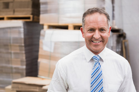 Senior man in business attire standing in warehouse among cardboard boxes and pallets, copy space. Industry, logistics, distribution, warehouse, shipping, storage, shipmentの写真素材