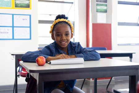African American girl sitting at classroom desk with notebook, red apple, posters, window blinds. Education, learning, student, study, academic, curriculum, schoolの写真素材