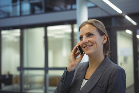 30s businesswoman wearing grey suit standing in office hallway speaking on smartphone, copy space. Professional, corporate, modern, communication, workplace, sleek, productivityの写真素材