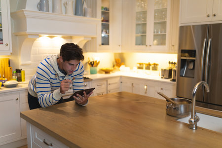 Middle-aged man leaning on kitchen island using tablet while stirring saucepan with wooden spoon. Modern, lifestyle, minimalism, technology, domestic, interior, relaxationの写真素材