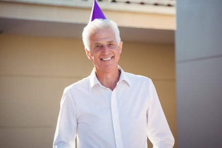 Senior man standing in driveway wearing purple party hat and white button-up shirt. Celebration, joy, elderly, outdoor, leisure, cheerful, lifestyleの写真素材