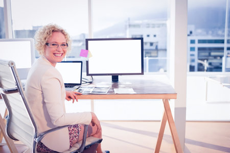 Woman sitting on office chair turning toward camera at wooden desk with monitor laptop, copy space. Professional, modern, minimalist, workplace, business, corporate, creativeの写真素材