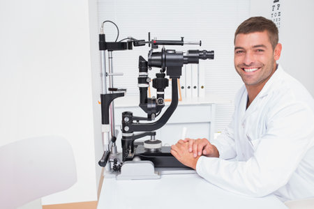 Male optometrist wearing lab coat sitting beside slit lamp biomicroscope at clinic desk, copy space....