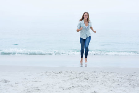 Woman running barefoot along wet sand at calm sea edge, wearing denim shirt and jeans. Carefree, adventure, serenity, casual, outdoor, lifestyle, fashionの写真素材