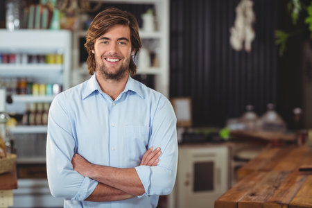 Bearded man standing with arms crossed in cafe with wooden service counter and glass cake domes. Coffeehouse, hospitality, casual, modern, interior, employer, decorの写真素材