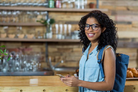 African American woman in 20s standing at cafe counter holding smartphone and backpack, copy space. Cafe, rustic, modern, casual, lifestyle, cozy, communityの写真素材