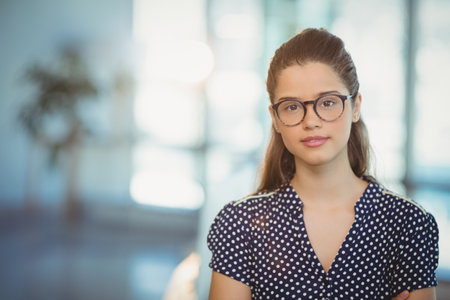 Woman wearing eyeglasses and navy polka-dot blouse standing in coworking space with potted plant. Professional, workspace, modern, natural light, elegant, focused, startupの写真素材