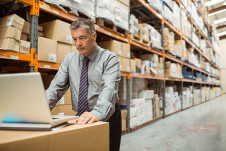Man in his 50s wearing tie typing on laptop at box in warehouse aisle, copy space. Industrial, logistics, professional, productivity, organization, workspace, modernの写真素材