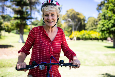 Senior woman holding bicycle handlebars in grassy park wearing red blouse, bicycle helmet. Activewear, outdoor recreation, vibrant, health, leisure, nature, lifestyleの写真素材