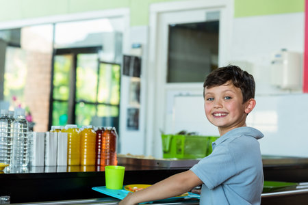 Smiling school-age boy holding lunch tray at school cafeteria counter by juice bottles, copy space. Children, education, nutrition, cheerful, bright, healthy, innocenceの写真素材
