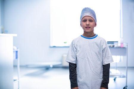 Boy standing in treatment room wearing surgical cap and gown by window and cart, copy space. Medical, healthcare, clinical, pediatrics, cleanliness, examination, hospitalの写真素材