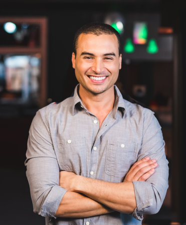 Man standing with arms crossed in softly lit lounge space with green pendant lights and shelves. Modern, professional, urban, cozy, ambient, stylish, masculineの写真素材