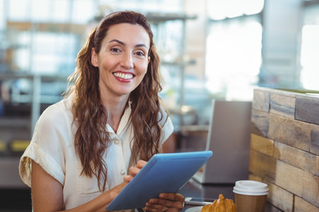 Woman smiling and holding tablet at cafe counter with coffee cup, croissant, copy space. Cafe,...