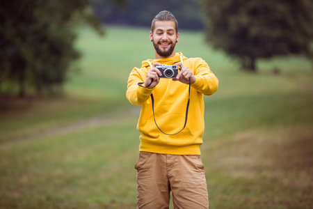 Man standing in grassy field wearing yellow hoodie and brown pants holding vintage camera....