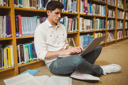 Male student typing on laptop while sitting on library floor with open textbook and notebook....