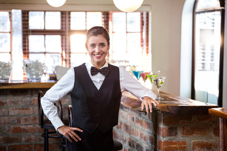Female bartender wearing uniform serving at brick counter offering colorful cocktails. Rustic,...