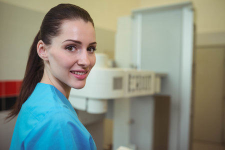 Female radiology technologist in scrubs looking toward camera at X-ray machine in lab, copy space. Medical, healthcare, diagnostic, professional, clinical, imaging, technologyの写真素材
